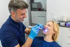 Dentist in blue gloves uses a dental curing light on a smiling patient in a clinic