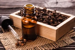 A small amber glass bottle with a dropper, next to a wooden box of dried cloves, rests on woven fabric