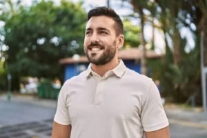 A smiling man with a beard wears a light polo shirt, standing outdoors