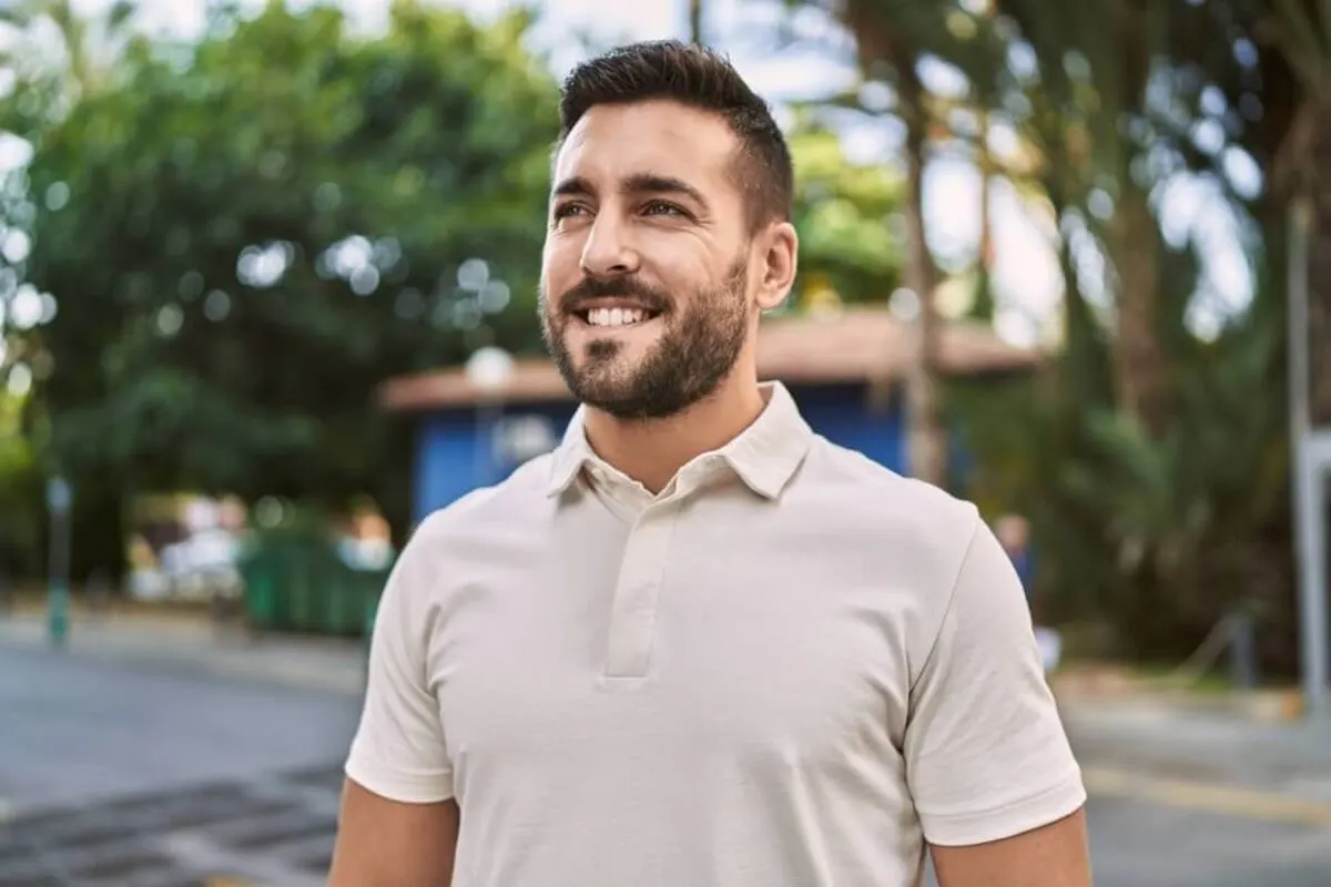 A smiling man with a beard wears a light polo shirt, standing outdoors