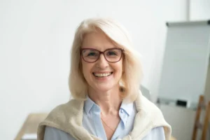 Smiling woman with glasses and shoulder-length blonde hair, wearing a light blue shirt and a sweater draped over her shoulders, in an office setting