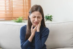 A woman in a blue sweater sits on a couch, grimacing and holding her cheek in pain, suggesting a toothache