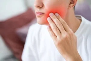 A person in a white shirt touches their cheek with a pained expression. The cheek is visibly red, suggesting toothache or facial pain