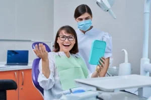 A joyful woman in a dental chair holds a hand mirror, smiling widely. A dentist stands beside her, masked, in a modern, well-lit dental office