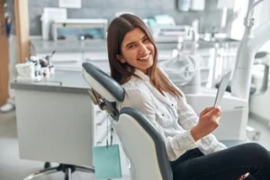 Smiling woman in a dental clinic sits on a treatment chair holding a hand mirror. The setting appears modern and clean, conveying a positive and relaxed atmosphere