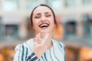 Woman in a blurred indoor setting, laughing joyfully while holding a clear dental aligner