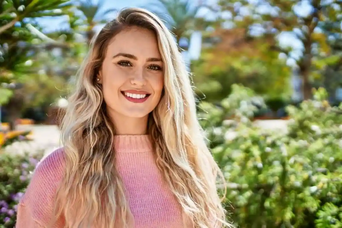 Smiling woman with long blonde hair in a pink sweater, standing in a sunny garden. Greenery and a clear blue sky create a cheerful, vibrant atmosphere.
