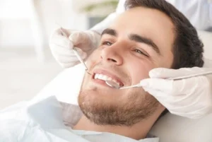 A man smiles in a dental chair while a dentist examines his teeth with tools. The scene conveys a sense of comfort and professionalism.