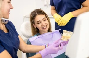 A woman smiles in a dental chair while a dentist in purple gloves shows a dental model
