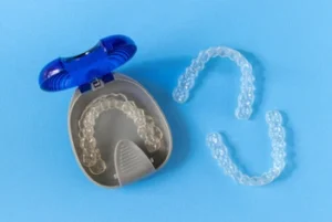 A blue retainer case is open, displaying a clear dental retainer inside. Two additional clear retainers are positioned nearby on a light blue background