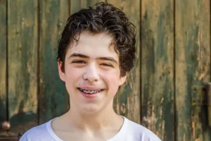 Smiling teenage boy with braces, wearing a white shirt. He's in front of a weathered, green wooden door, conveying a cheerful and relaxed vibe
