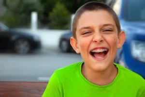A young boy with short hair and braces laughs joyfully, wearing a bright green shirt