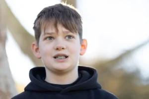 A child with short brown hair and braces, wearing a black hoodie, stands outdoors