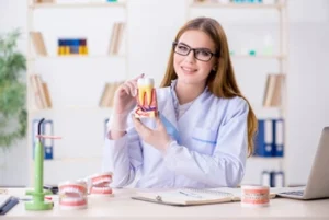 Full Mouth Dental Implants Cost australia explanation A dentist, wearing glasses and a white coat, smiles while holding a dental model in an office