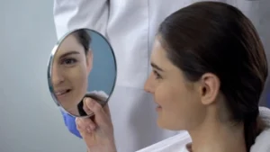 A woman with dark hair smiles as she looks at her reflection in a handheld mirror. She's in a medical setting, suggesting a positive outcome.