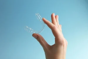 A hand holds a clear dental aligner against a light blue background. The image conveys a clean, minimalistic feel, highlighting oral care and orthodontics
