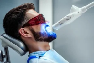 Man undergoing teeth whitening, wearing protective glasses, reclined in a dental chair. A bright blue light is directed at his mouth