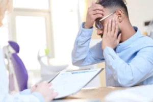 sinus tooth pain symptoms A worried man in glasses holds his head, sitting across from someone holding a clipboard