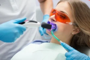 how long does teeth whitening last professional A woman receives a dental treatment, wearing orange protective glasses. A dentist uses a tool with blue light on her mouth