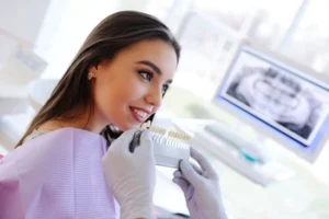 Teeth Whitening Bali procedure A smiling woman in a dental office wears a lilac bib, while a gloved dentist holds a tooth color chart to her teeth; an X-ray visible on a monitor