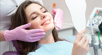 A female patient reclining in a dental chair, smiling as she examines her teeth in a hand mirror following a successful procedure performed by her dentist