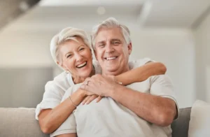 Elderly couple smiling warmly, woman embracing man from behind on a couch in a bright room