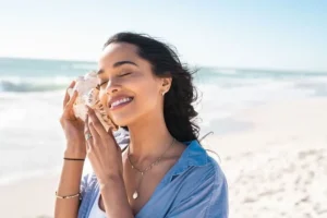 A woman holding a seashell to her ear to listen to the ocean while on the beach