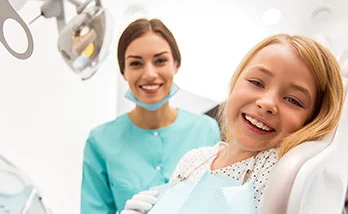 A young girl smiles while seated in a dental chair together with a dentist