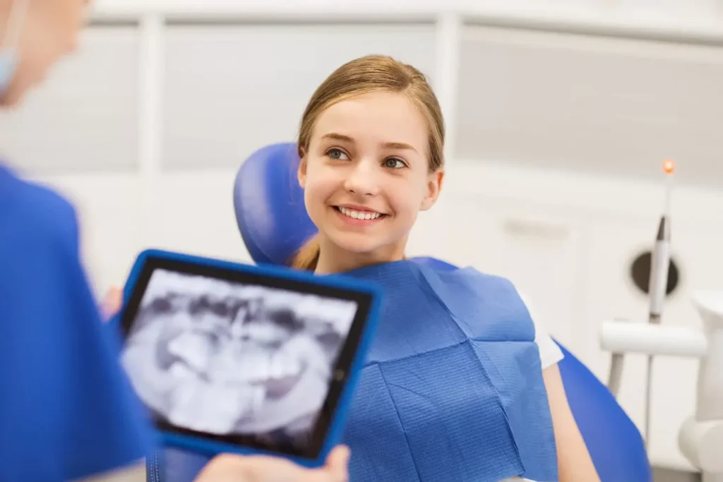 A young girl smiles sitting in a dental chair. A dentist holds a tablet displaying an X-ray