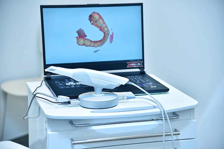 A dental scanner sits on a white cabinet beside a laptop displaying a 3D scan of teeth. The setting is bright and clinical, conveying a modern, professional tone