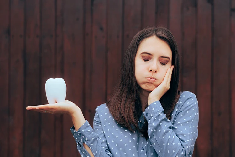 A woman holds her cheek, appearing in pain, with a large tooth model on her palm