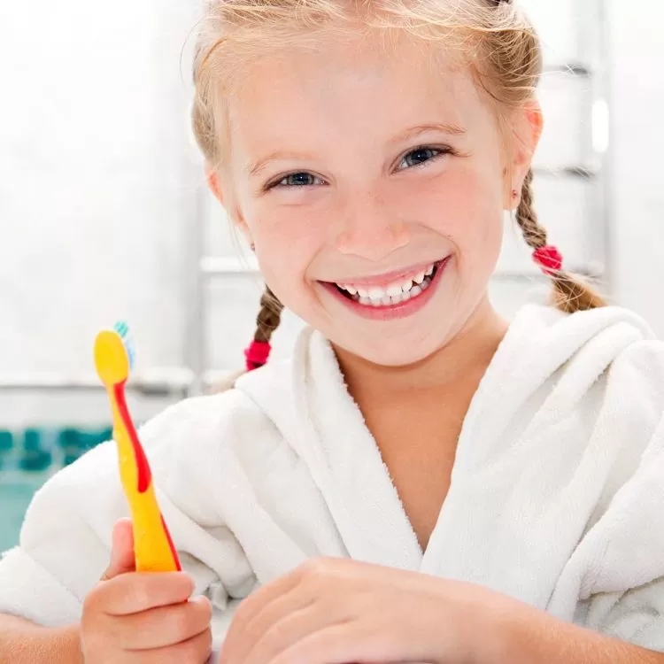 Smiling girl with braids in a white robe holds a colorful toothbrush, standing in a bright bathroom, conveying a cheerful and playful morning routine.
