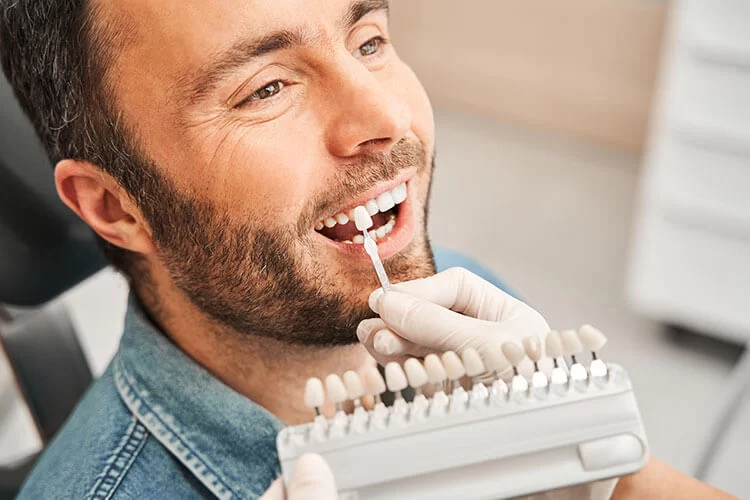 A smiling man in a dentist's chair is having his teeth color-matched with a shade guide