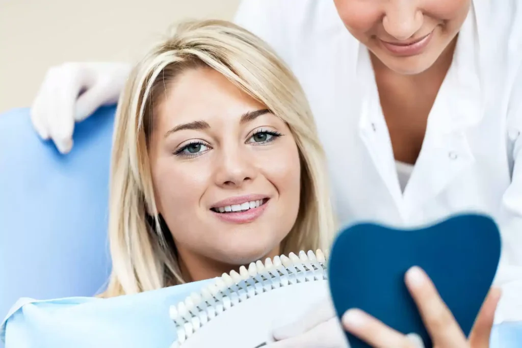 A woman smiling in a dental chair as she holds a tooth shade guide for teeth color selection