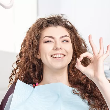 A smiling woman sits in a dental chair making an "OK" gesture with her hand