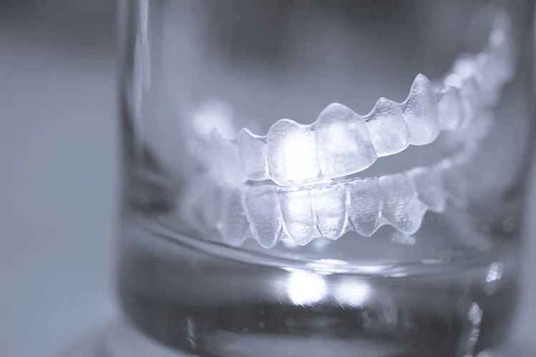 Clear dental aligners rest in a glass, reflecting on the surface. The transparent material highlights the teeth mold details, evoking cleanliness.