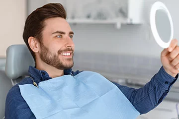 A man in a dental chair holding a mirror and smiling at his new, bright smile following a professional teeth-whitening treatment in a modern dental office.